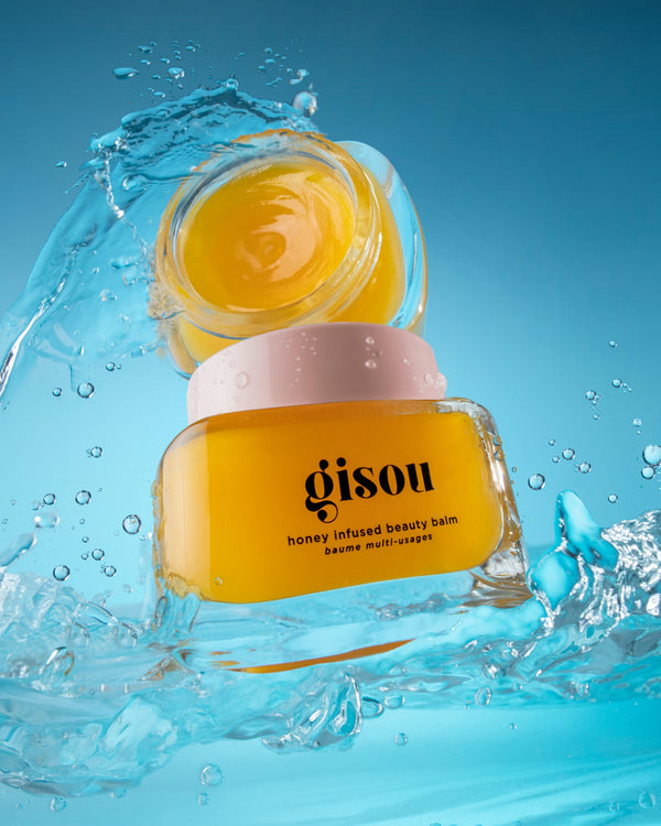 A closed jar of honey infused beauty balm next to an opened jar of beauty balm on the blue background with splashes of water